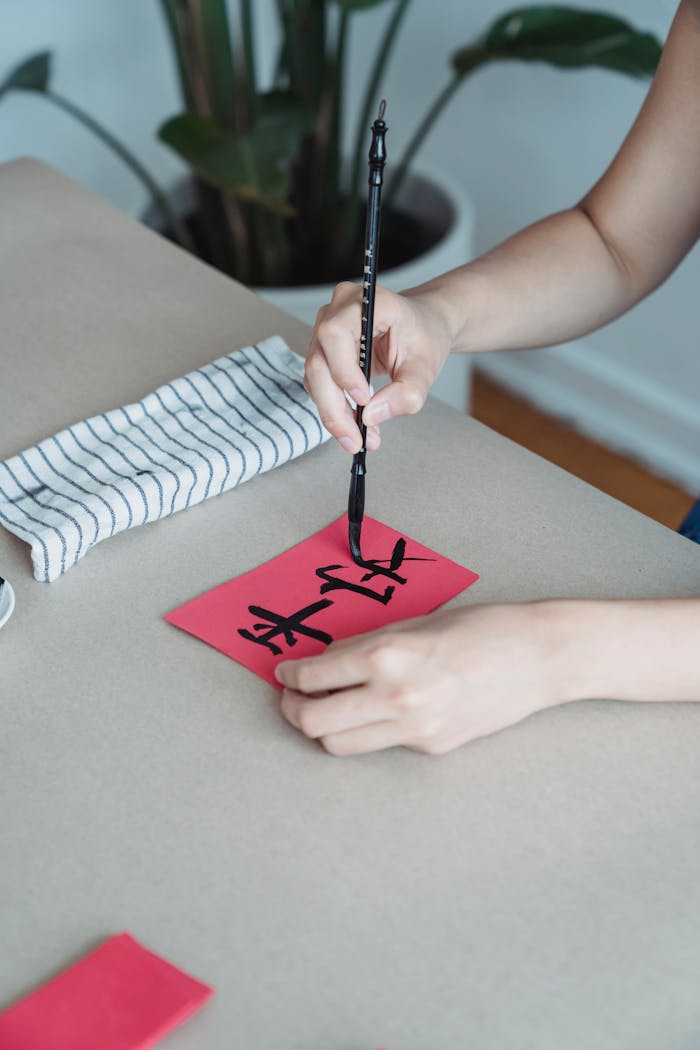 A person practices calligraphy on red paper using a brush pen on a white table.